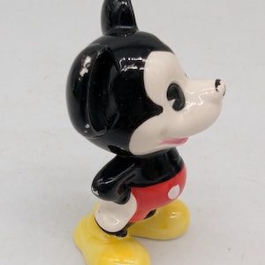May include: A vintage Mickey Mouse figurine. The figure has a black head and ears, a white face, red shorts with white buttons, and yellow shoes. The figurine is a collectible item.