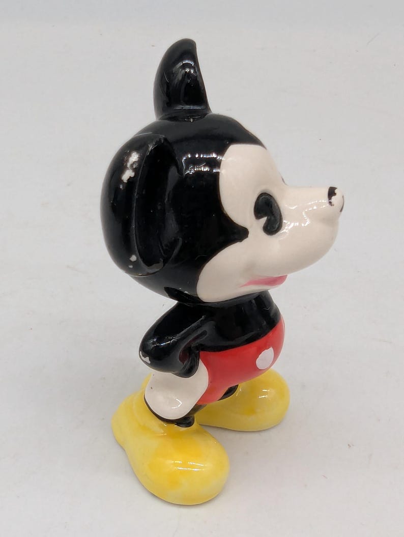 May include: A vintage Mickey Mouse figurine. The figure has a black head and ears, a white face, red shorts with white buttons, and yellow shoes. The figurine is a collectible item.