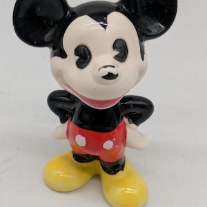 May include: A ceramic Mickey Mouse figurine. The figure has a black head and ears, a white face, red shorts with white polka dots, yellow shoes, and white gloves. The figurine is standing with its arms at its sides.