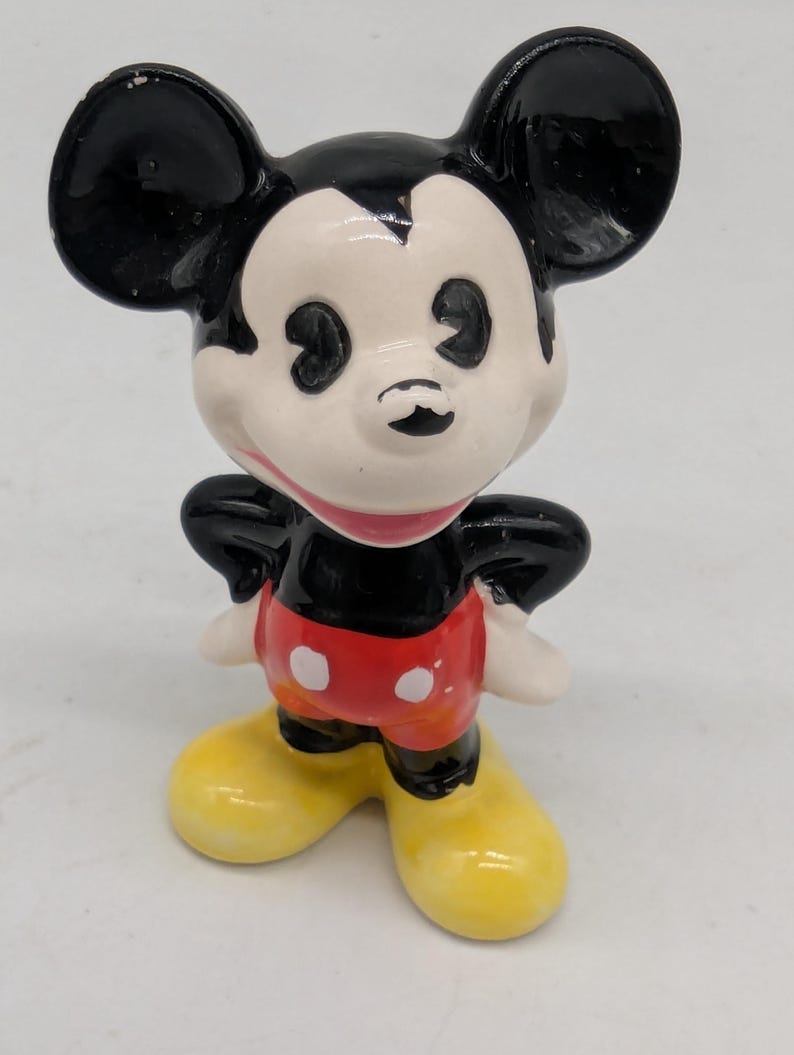 May include: A ceramic Mickey Mouse figurine. The figure has a black head and ears, a white face, red shorts with white polka dots, yellow shoes, and white gloves. The figurine is standing with its arms at its sides.