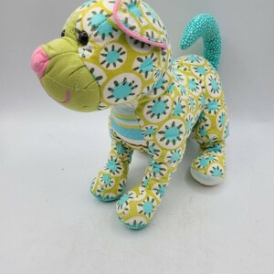 May include: A stuffed dog toy with a patterned design. The dog's body is covered in a yellow fabric with blue and white floral accents. The dog has a green snout, pink nose, and a blue and white striped collar.