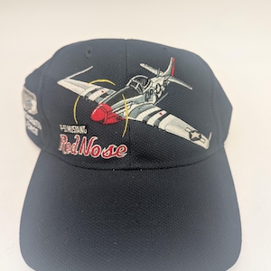 May include: A navy blue baseball cap with an embroidered P-51 Mustang fighter plane design. The plane has a red nose and is set against a grey and white background. The text "P-51 Mustang Red Nose" is embroidered below the plane. The cap has a curved peak.