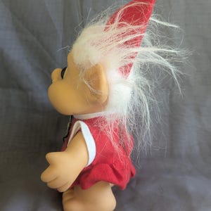 Puede incluir: Una mu&ntilde;eca troll vintage con un vestido rojo con ribete blanco y un sombrero rojo a juego con un pomp&oacute;n blanco. La mu&ntilde;eca tiene pelo blanco y esponjoso y una expresi&oacute;n sonriente. El troll es un art&iacute;culo de colecci&oacute;n.