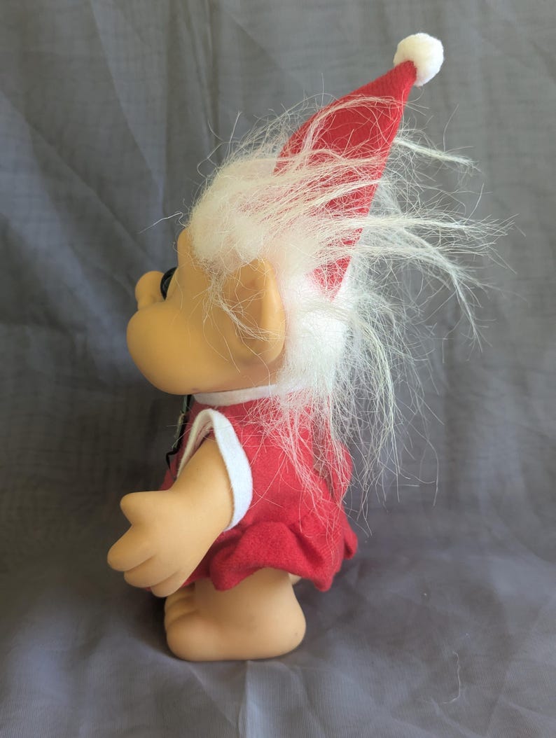 Puede incluir: Una mu&ntilde;eca troll vintage con un vestido rojo con ribete blanco y un sombrero rojo a juego con un pomp&oacute;n blanco. La mu&ntilde;eca tiene pelo blanco y esponjoso y una expresi&oacute;n sonriente. El troll es un art&iacute;culo de colecci&oacute;n.