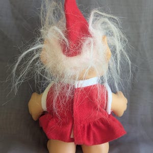 Puede incluir: Una mu&ntilde;eca troll vintage, vista desde atr&aacute;s, con un vestido rojo y un gorro de Pap&aacute; Noel con un pomp&oacute;n blanco. La mu&ntilde;eca tiene pelo largo, blanco y desgre&ntilde;ado y una cara bronceada. Un ribete blanco adorna el vestido.