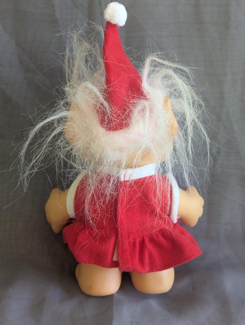 Puede incluir: Una mu&ntilde;eca troll vintage, vista desde atr&aacute;s, con un vestido rojo y un gorro de Pap&aacute; Noel con un pomp&oacute;n blanco. La mu&ntilde;eca tiene pelo largo, blanco y desgre&ntilde;ado y una cara bronceada. Un ribete blanco adorna el vestido.
