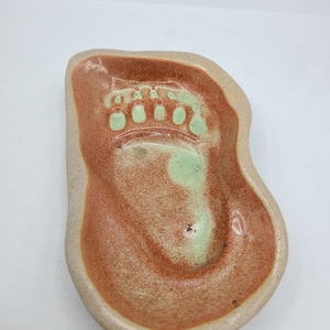 May include: A ceramic foot-shaped dish with a light brown rim and a reddish-brown interior. The interior features a green foot imprint. The dish is designed to hold small items.