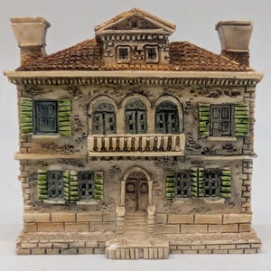 May include: A detailed miniature house figurine with a textured, stone-like facade. The model features a brown tiled roof, green shutters, and a small balcony. The house has two chimneys and a front door with steps.