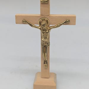 May include: A wooden crucifix with a gold-colored figure of Jesus Christ. The cross is made of light-colored wood, with a small plaque at the top. The figure is detailed, with arms outstretched. The base is also made of wood.