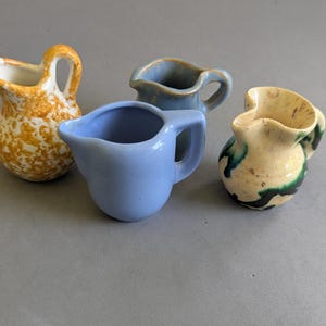May include: A collection of four small ceramic jugs. One is yellow with a white speckled pattern, one is solid light blue, one is a muted blue-grey, and one is cream-coloured with dark green and black abstract painted accents.