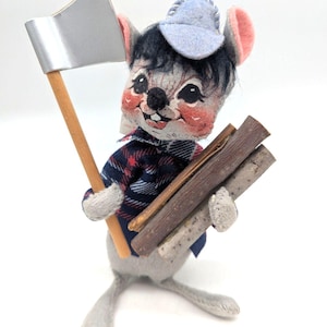 May include: A whimsical, handmade mouse figurine dressed as a lumberjack. The mouse wears a blue plaid shirt, a light blue cap, and carries a silver axe and a bundle of wooden sticks. The mouse has rosy cheeks and a cheerful expression.