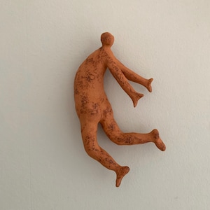 Pode incluir: Uma escultura de parede na cor terracota representando uma figura humana estilizada numa pose dinâmica de salto. A figura tem uma superfície texturizada e manchada com pontos castanhos escuros, cabeça arredondada e membros estendidos em movimento.