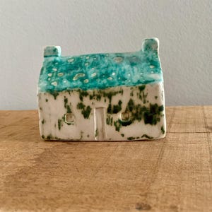 Op de afbeelding: Een klein keramisch huisje met een turquoise dak en witte muren. Het dak heeft een gespikkeld patroon en de muren zijn versierd met groene vlekken. Het huis heeft een deur en twee kleine ramen. Het is een decoratief item.