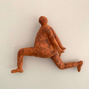 Peut inclure: Sculpture en céramique d'une personne en mouvement, courant. La figurine orange est parsemée de taches brun foncé, fixée sur un mur blanc. Les bras et les jambes sont tendus, suggérant le mouvement.