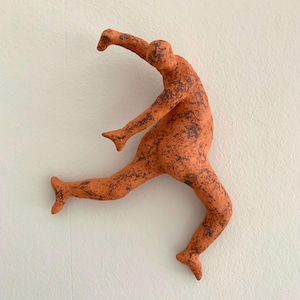 Può includere: Scultura in ceramica arancione di una persona in posa di corsa. La figura ha una superficie strutturata con macchie marrone scuro. La scultura è montata su una parete bianca.