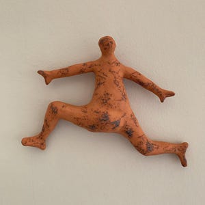 Könnte beinhalten: Eine terrakottafarbene Wandskulptur, die eine stilisierte menschliche Figur in einer dynamischen Sprungpose darstellt. Die Oberfläche der Figur weist ein gesprenkeltes, dunkelbraun texturiertes Finish an Armen, Rumpf und Beinen auf.