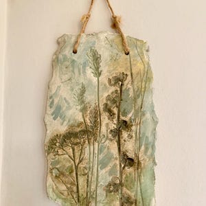 May include: A rectangular ceramic wall hanging with a natural, textured edge. The artwork features a painted landscape with tall, slender plants in shades of green, brown, and beige against a light blue and cream background. A jute rope is attached for hanging.