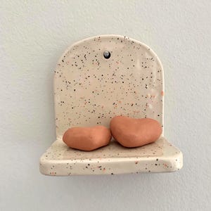 May include: A speckled ceramic shelf with two heart-shaped objects. The shelf is off-white with black and orange speckles. The hearts are a muted orange color. The shelf has a rounded top and a small hole for hanging.