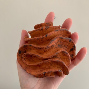 Op de afbeelding: Een handgemaakte, oranje keramische roos sculptuur met donkerblauwe accenten. De bloem wordt in een open hand gehouden en toont zijn getextureerde, gelaagde bloemblaadjes. De kleur en het ontwerp van de sculptuur suggereren een rustiek of artistiek thema.
