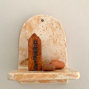 Può includere: Una mensola da parete in ceramica a forma di arco con finitura maculata color crema e marrone chiaro. Sulla mensola poggia un segnaposto in argilla arancione a punta con testo in rilievo scuro e due pietre marroni lisce e arrotondate.