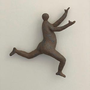 May include: A brown, textured wall sculpture depicting a stylised human figure in a dynamic, leaping pose. The figure has a rounded torso, outstretched arms reaching upward, and legs extended in a mid-air motion, finished with a matte, earthy surface.