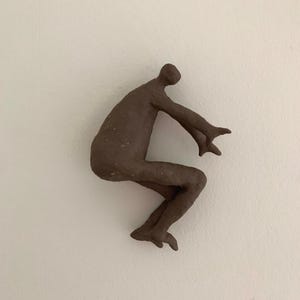 Op de afbeelding: Een donkerbruin, getextureerd kleibeeld van een menselijke figuur in een gehurkte, springende houding. Het figuur heeft een afgerond hoofd, een gebogen rug, gebogen knieën en armen die naar voren zijn gestrekt met open handen.