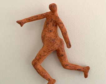 May include: A terracotta-colored ceramic wall sculpture depicting a stylized human figure in a dynamic, running pose. The surface features a textured, mottled finish with dark brown speckles throughout the clay body, showing one arm extended forward and legs positioned in mid-stride.
