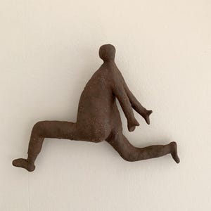 Puede incluir: Una escultura de pared de cerámica marrón y texturizada que representa una figura humana estilizada en una pose dinámica de carrera o salto. La figura tiene una cabeza redondeada, un torso, dos brazos extendidos hacia adelante y dos piernas en movimiento.