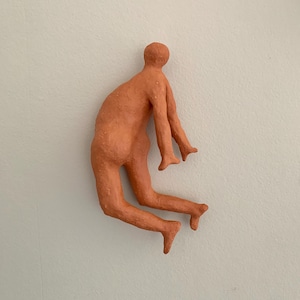 Può includere: Una scultura color terracotta di una figura umana in una posa dinamica, che sembra essere in movimento. La figura è fissata a un muro bianco, mostrando la consistenza e la forma. Le braccia e le gambe sono estese, trasmettendo un senso di movimento.