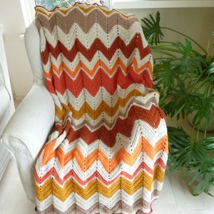 Può includere: Coperta all'uncinetto fatta a mano con motivo a zig-zag nei toni crema, marrone, arancione e giallo, drappeggiata su una poltrona bianca. Un elemento d'arredo caldo e accogliente.