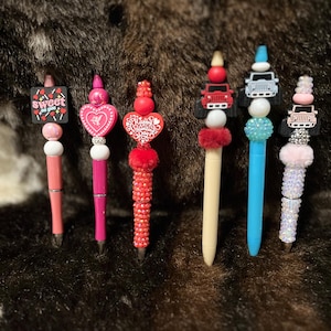 May include: Six decorative pens with various embellishments. The pens feature designs such as a heart, a Jeep, and the words "Sweet on you" and "Happy Valentine's Day." The pens are in shades of pink, red, cream, and blue.