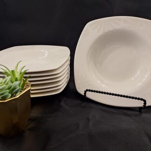 May include: A set of white square ceramic bowls with rounded corners and a subtle embossed floral pattern on the wide rim. One bowl is displayed upright on a black wire stand, while a stack of six identical bowls sits to the left.