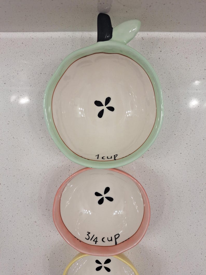Pu&ograve; includere: Set di tre misurini in ceramica a forma di mela. La tazza superiore &egrave; verde e contrassegnata con "1 cup". La tazza centrale &egrave; rosa e contrassegnata con "3/4 cup". Ogni tazza presenta un motivo floreale nero al centro.