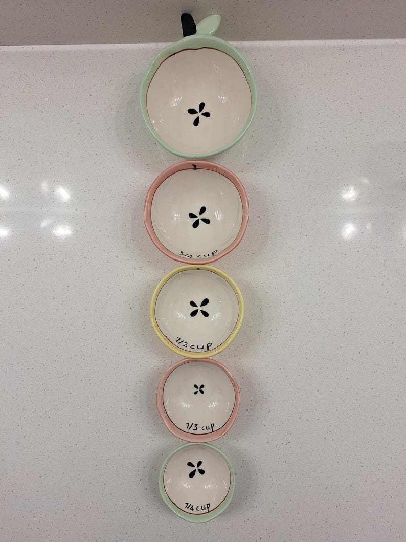 Pu&ograve; includere: Un set di quattro ciotole dosatrici in ceramica a forma di mela, impilate verticalmente. Le ciotole sono bianche con bordi color pastello e dettagli di semi neri. Ogni ciotola &egrave; etichettata con una frazione di tazza.