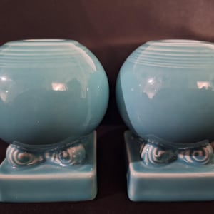 May include: Two turquoise ceramic vases with a spherical shape and a square base. Each vase has a series of white stripes around the top and decorative swirls on the base. The vases are displayed on a dark background.