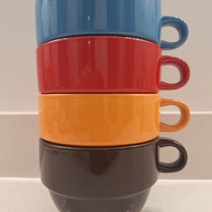 May include: A stack of four ceramic soup bowls in blue, red, orange, and brown. Each bowl has a rounded shape with a handle. The bowls are stacked on top of each other, showcasing their vibrant colors and design.
