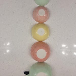 Pu&ograve; includere: Cinque ciotole in ceramica dai colori pastello disposte verticalmente: verde, rosa e giallo. Le ciotole sono rotonde con interno bianco. La ciotola inferiore ha un manico e il testo "ELISE LEFEBVRE CERAMIQUE".
