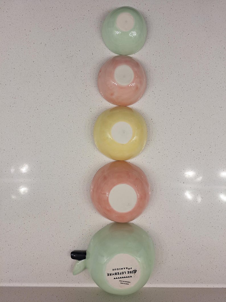 Pu&ograve; includere: Cinque ciotole in ceramica dai colori pastello disposte verticalmente: verde, rosa e giallo. Le ciotole sono rotonde con interno bianco. La ciotola inferiore ha un manico e il testo "ELISE LEFEBVRE CERAMIQUE".