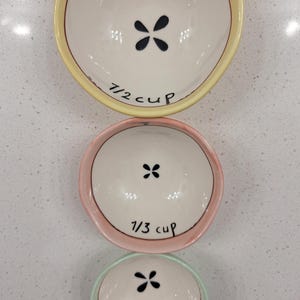 Pu&ograve; includere: Tre misurini in ceramica disposti verticalmente. La tazza superiore &egrave; gialla, con la scritta "1/2 cup" e un motivo floreale nero. La tazza centrale &egrave; rosa, con la scritta "1/3 cup". La tazza inferiore &egrave; verde, con la scritta "1/4 cup".