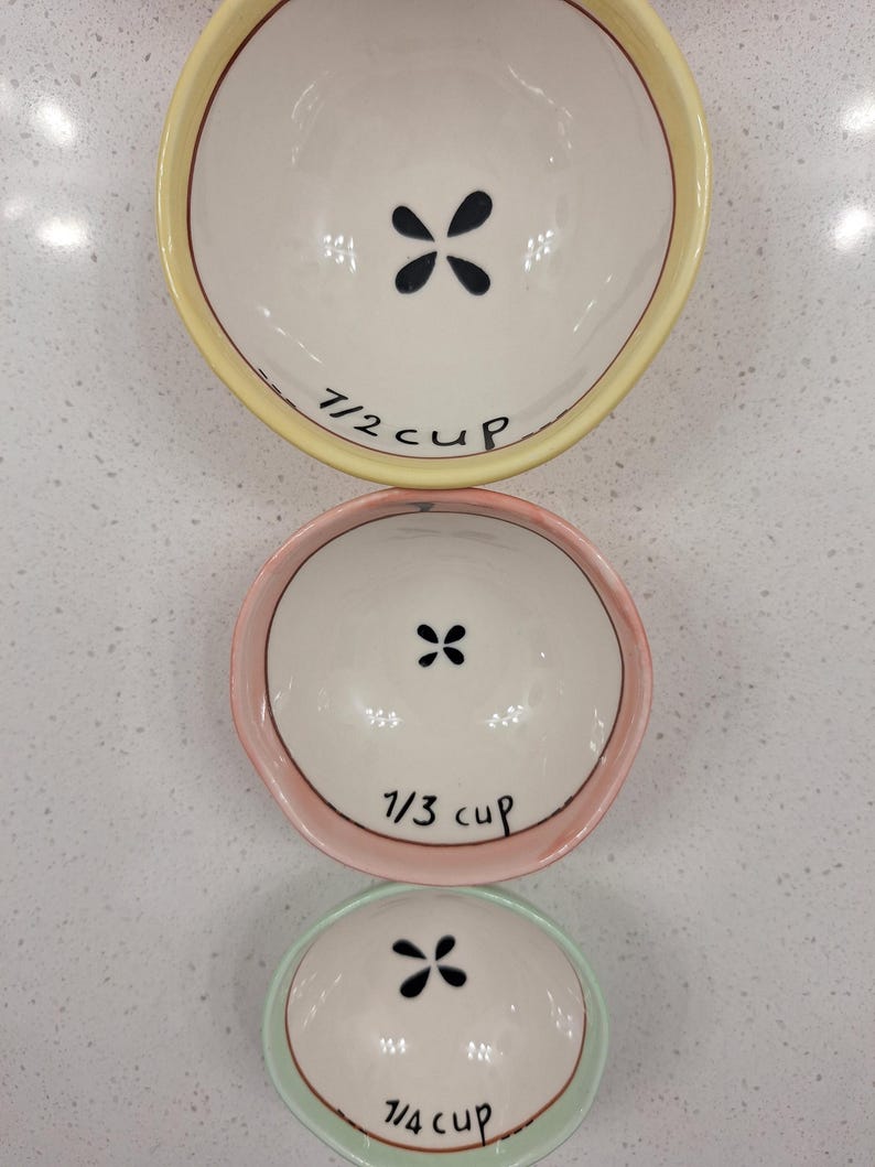 Pu&ograve; includere: Tre misurini in ceramica disposti verticalmente. La tazza superiore &egrave; gialla, con la scritta "1/2 cup" e un motivo floreale nero. La tazza centrale &egrave; rosa, con la scritta "1/3 cup". La tazza inferiore &egrave; verde, con la scritta "1/4 cup".