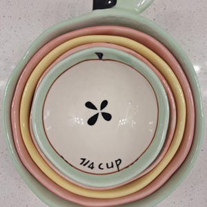 Pu&ograve; includere: Un set di misurini in ceramica a forma di mela, nei colori pastello verde, rosa e giallo. La tazza pi&ugrave; piccola &egrave; bianca con un disegno floreale nero e la scritta "7/4 cup".