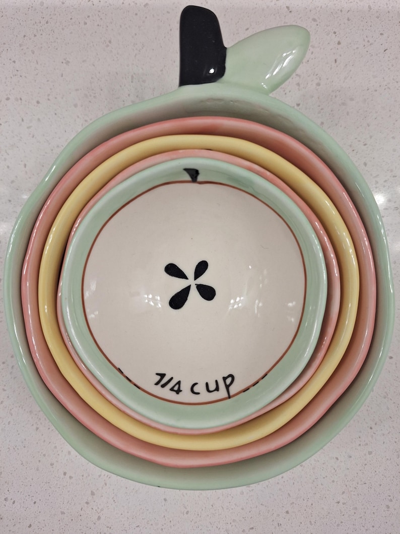 Pu&ograve; includere: Un set di misurini in ceramica a forma di mela, nei colori pastello verde, rosa e giallo. La tazza pi&ugrave; piccola &egrave; bianca con un disegno floreale nero e la scritta "7/4 cup".