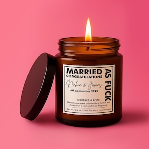 May include: A lit candle in an amber glass jar with a black lid. The white label reads MARRIED AS FUCK CONGRATULATIONS Nickie & James 6th September 2025 RHUBARB & ROSE Delicate rose scent balanced by a hint of tart rhubarb 245 ml / 8.6 oz 100% Soy Wax.