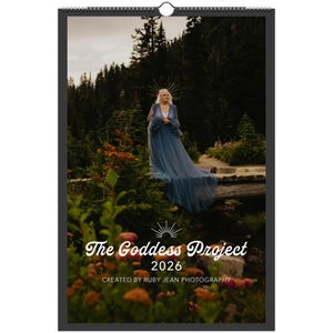 May include: A calendar featuring a woman in a flowing blue gown, standing in a forest setting. The calendar is titled "The Goddess Project 2026" with the photographer's name below. The image has a natural, ethereal aesthetic, with a focus on the woman and the surrounding landscape.