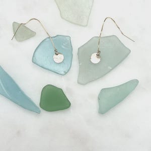May include: Silver threader earrings with hammered disc charms. The earrings are displayed on a white surface with pieces of sea glass in shades of blue, green, and white.
