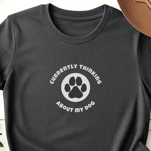Könnte beinhalten: Dunkelgraues T-Shirt mit umgekrempelten Ärmeln. Das Shirt zeigt eine weiße kreisförmige Grafik mit einem Hundepfotenabdruck und dem Text "CURRENTLY THINKING ABOUT MY DOG". Das T-Shirt besteht aus einem weichen Material.