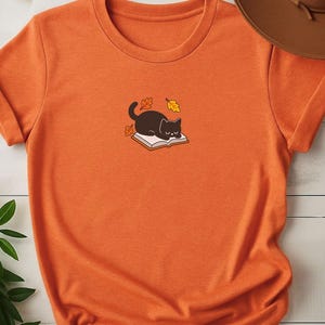 Op de afbeelding: Een heather oranje t-shirt met korte mouwen en een centrale afbeelding van een zwarte kat die op een open boek slaapt, omringd door kleine oranje en gele herfstbladeren. Het shirt heeft een ronde hals en een eenvoudig, strak ontwerp.