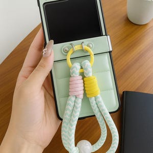 Puede incluir: Una funda de teléfono verde menta con borde negro y diseño acolchado. Cuenta con un anillo amarillo y una correa de cuerda con detalles en rosa, amarillo y blanco. La funda del teléfono es sostenida por una mano.