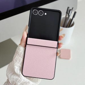 May include: A pink, leather-effect phone case with a black frame, open to show a black interior and camera lenses. The case is held up, displaying its design and texture. A small pink square is visible in the background.