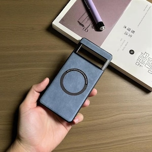 May include: A blue phone case with a magnetic card holder. The case features a rectangular camera cutout and a circular magnetic ring. The text "KEEP WITH MOBILE MAGNETIC CARD HOLDER" is printed on the case. A book and pen are in the background.